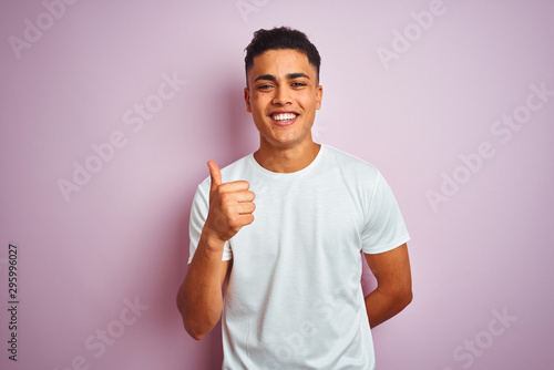 Young brazilian man wearing t-shirt standing over isolated pink background doing happy thumbs up gesture with hand. Approving expression looking at the camera with showing success.