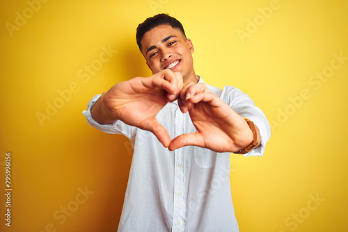 Young brazilian businessman wearing elegant shirt standing over isolated yellow background smiling in love showing heart symbol and shape with hands. Romantic concept.