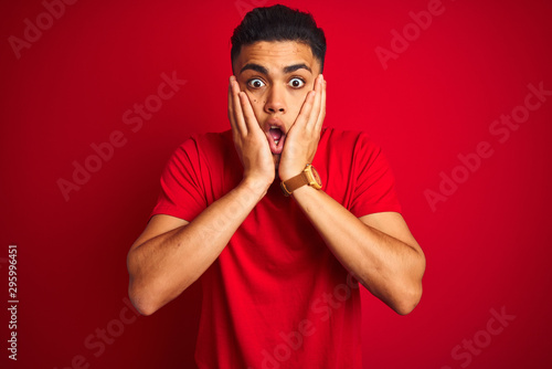 Young brazilian man wearing t-shirt standing over isolated red background afraid and shocked, surprise and amazed expression with hands on face