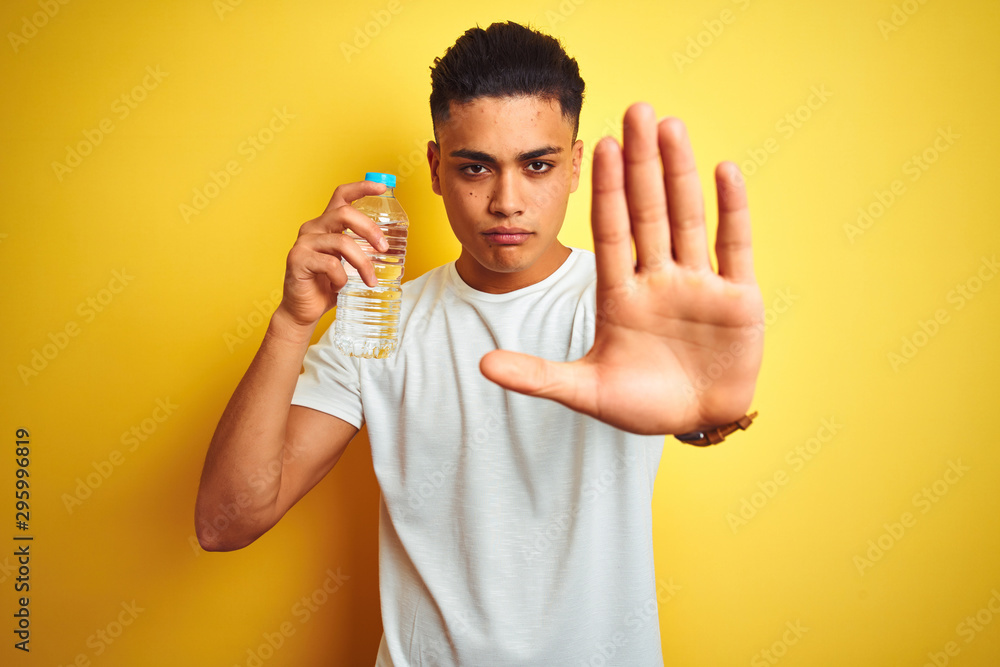 Young brazilian man holding bottle of water standing over isolated yellow background with open hand doing stop sign with serious and confident expression, defense gesture Young brazilian man holding bottle of water standing over isolated yellow background with open hand doing stop sign with serious and confident expression, defense gesture