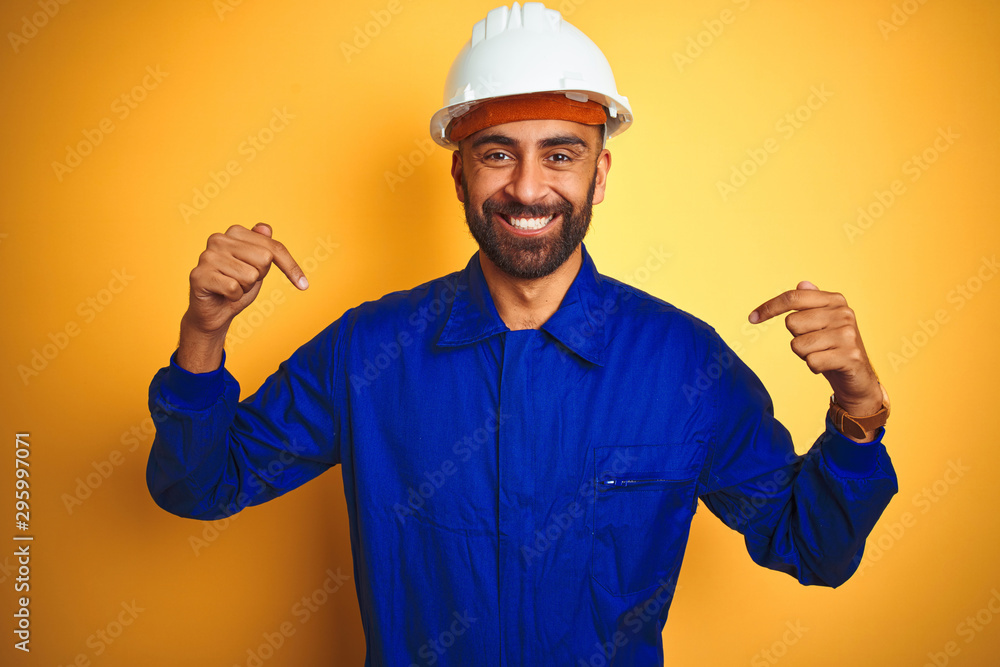 Handsome indian worker man wearing uniform and helmet over isolated yellow background looking confident with smile on face, pointing oneself with fingers proud and happy.