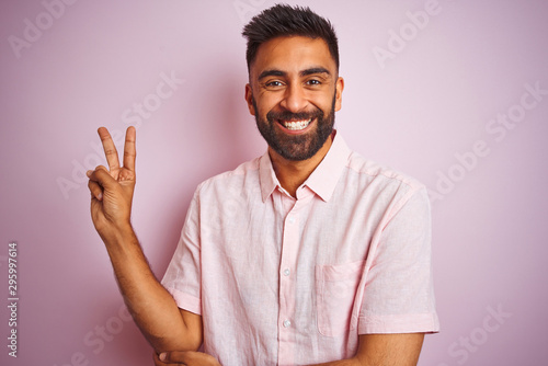 Young indian man wearing casual shirt standing over isolated pink background smiling with happy face winking at the camera doing victory sign. Number two.