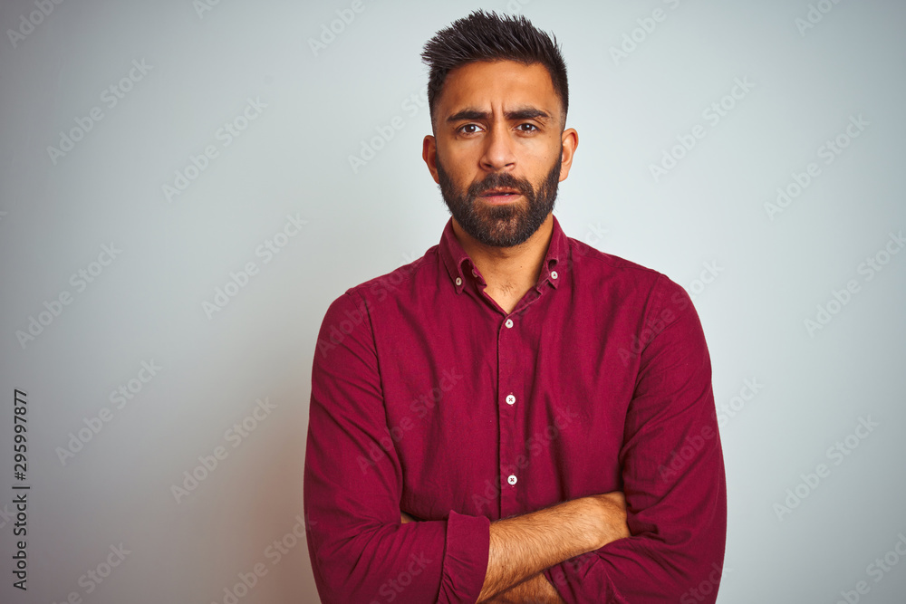 Young indian man wearing red elegant shirt standing over isolated grey background skeptic and nervous, disapproving expression on face with crossed arms. Negative person.
