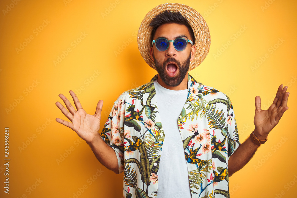 Indian man on vacation wearing floral shirt hat sunglasses over isolated yellow background celebrating crazy and amazed for success with arms raised and open eyes screaming excited. Winner concept