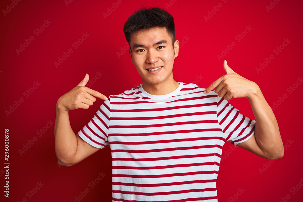 Young asian chinese man wearing striped t-shirt standing over isolated red background looking confident with smile on face, pointing oneself with fingers proud and happy. Young asian chinese man wearing striped t-shirt standing over isolated red background looking confident with smile on face, pointing oneself with fingers proud and happy.