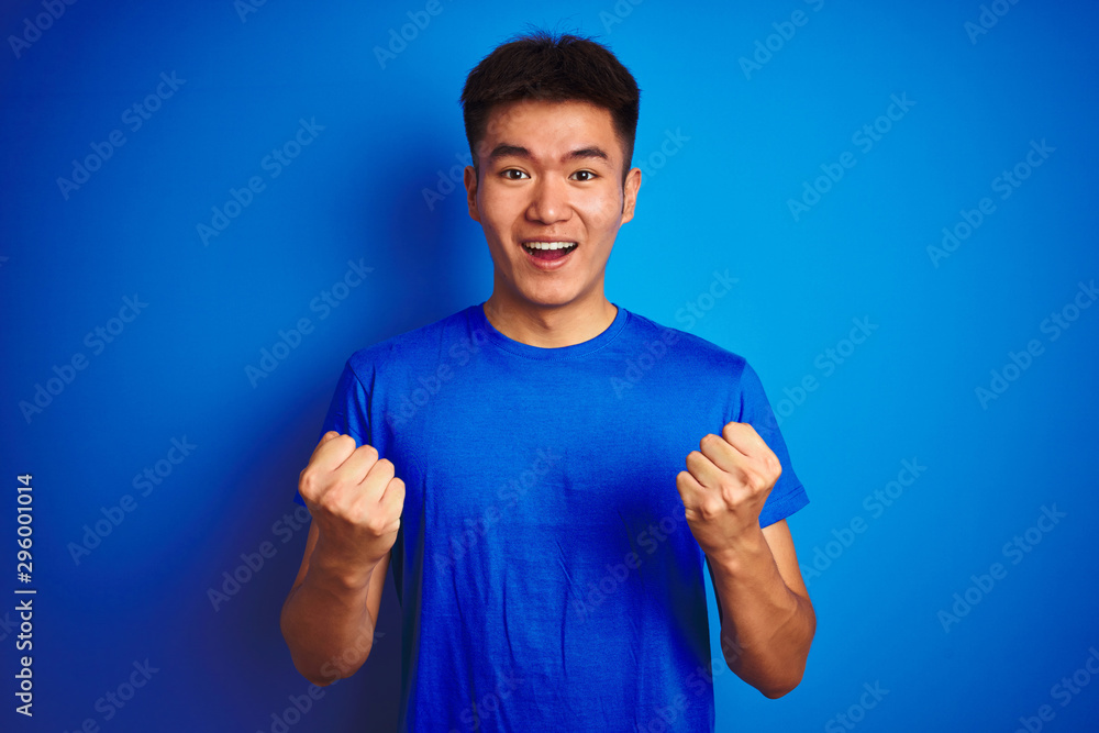 Fototapeta premium Young asian chinese man wearing t-shirt standing over isolated blue background celebrating surprised and amazed for success with arms raised and open eyes. Winner concept.