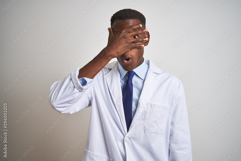 Young african american doctor man wearing coat standing over isolated white background peeking in shock covering face and eyes with hand, looking through fingers with embarrassed expression.