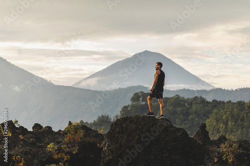 Young man standing on top of stone and enjoying amazing volcanic mountain Agung view in Bali. Travel and active lifestyle concept. photo