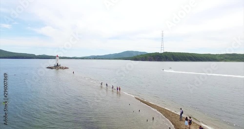 Flying above sand bar leading to the Tokarevsky lighthouse in Vladivostok. People sunbathe and walk on water to the lighthouse. Morot boat. Summer day. Russia. Aerial photo