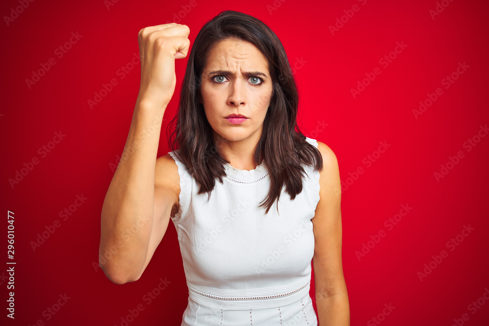 Young beautiful woman wearing white dress standing over red isolated background angry and mad raising fist frustrated and furious while shouting with anger. Rage and aggressive concept. Young beautiful woman wearing white dress standing over red isolated background angry and mad raising fist frustrated and furious while shouting with anger. Rage and aggressive concept.