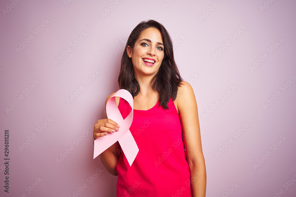 Young beautiful woman holding cancer ribbon over pink isolated background with a happy face standing and smiling with a confident smile showing teeth Young beautiful woman holding cancer ribbon over pink isolated background with a happy face standing and smiling with a confident smile showing teeth
