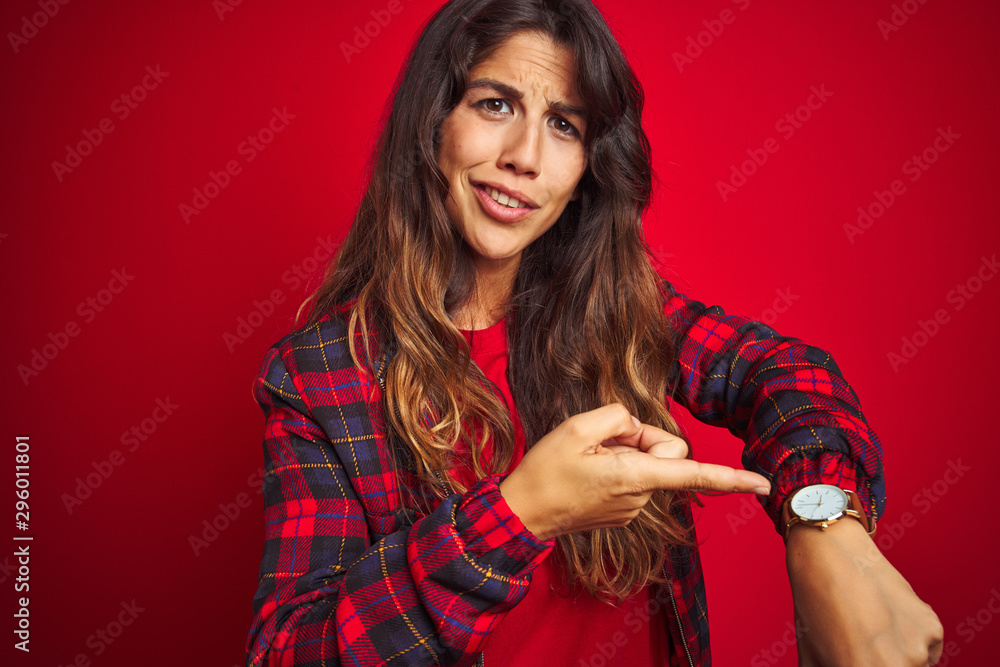 Young beautiful woman wearing casual jacket standing over red isolated background laughing at you, pointing finger to the camera with hand over mouth, shame expression