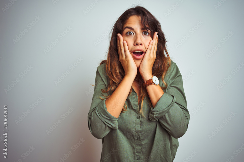 Young beautiful woman wearing green shirt standing over grey isolated background afraid and shocked, surprise and amazed expression with hands on face