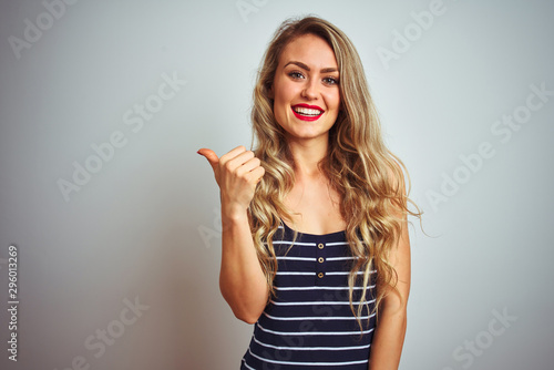 Young beautiful woman wearing stripes t-shirt standing over white isolated background smiling with happy face looking and pointing to the side with thumb up.
