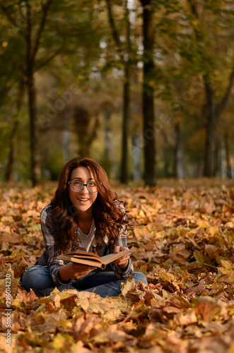 Beautiful girl in glasses sitting on the ground and reading book in park. Leisure time on warm autumn day. Autumn mood, enjoy the season