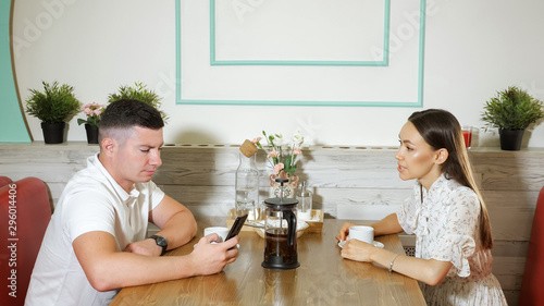 beautiful long haired girl talks with young guy using smartphone sitting at served table in cozy modern cafe