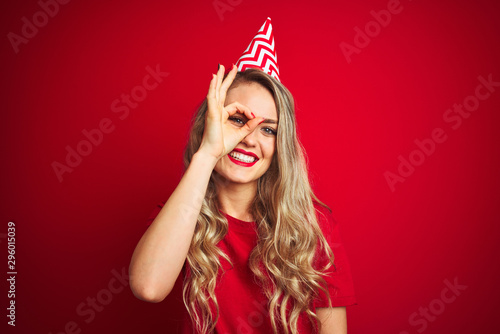 Young beautiful woman wearing bitrhday hat over red isolated background with happy face smiling doing ok sign with hand on eye looking through fingers
