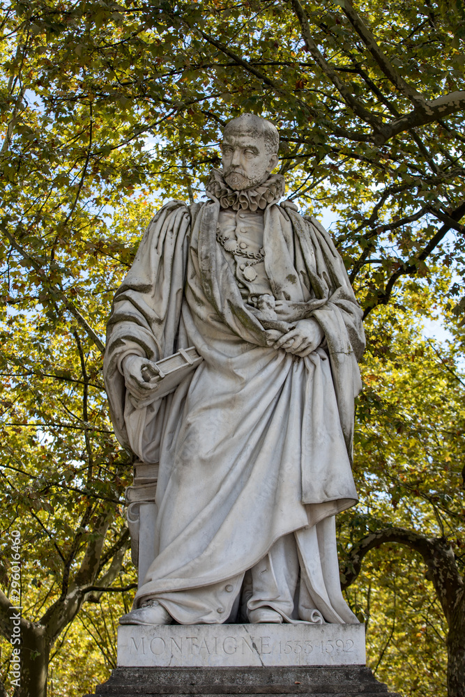 Statue of Michel de Montaigne in public garden along Place des Quinconces, Bordeaux France, with a canopy of green trees.