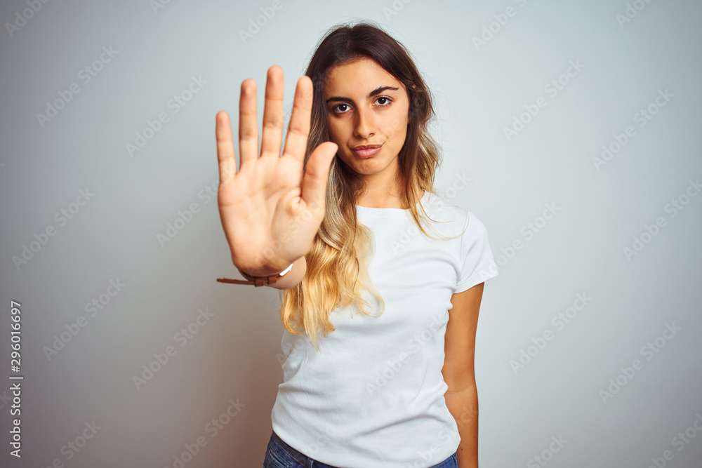 Young beautiful woman wearing casual white t-shirt over isolated background doing stop sing with palm of the hand. Warning expression with negative and serious gesture on the face. Young beautiful woman wearing casual white t-shirt over isolated background doing stop sing with palm of the hand. Warning expression with negative and serious gesture on the face.
