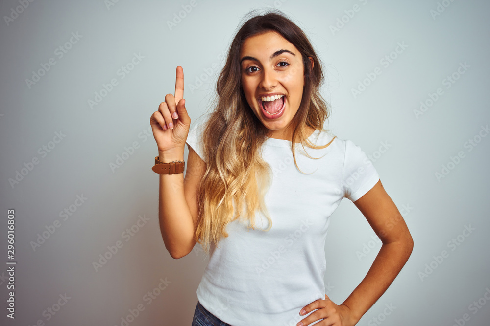 Young beautiful woman wearing casual white t-shirt over isolated background pointing finger up with successful idea. Exited and happy. Number one. Young beautiful woman wearing casual white t-shirt over isolated background pointing finger up with successful idea. Exited and happy. Number one.