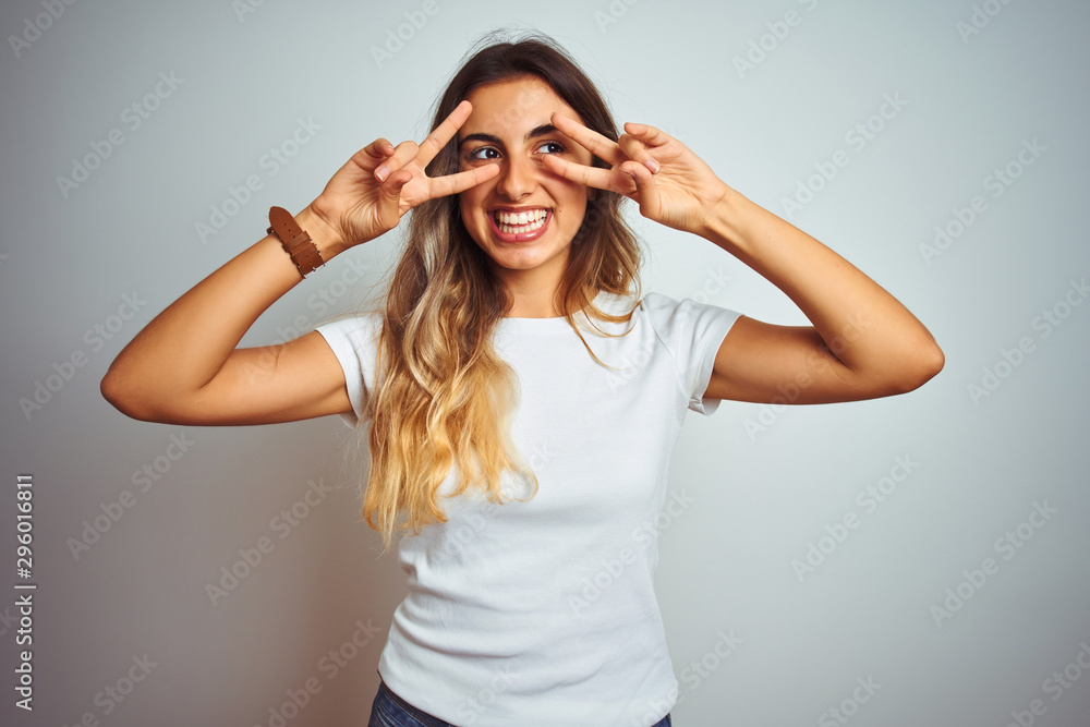 Naklejka premium Young beautiful woman wearing casual white t-shirt over isolated background Doing peace symbol with fingers over face, smiling cheerful showing victory