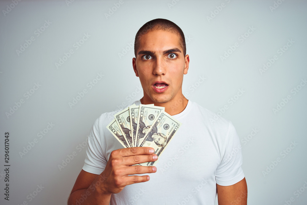 Young handsome man holding dollars over white isolated background scared in shock with a surprise face, afraid and excited with fear expression Young handsome man holding dollars over white isolated background scared in shock with a surprise face, afraid and excited with fear expression