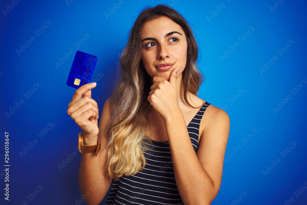 Young beautiful woman holding credit card over blue isolated background serious face thinking about question, very confused idea - obrazy, fototapety, plakaty 