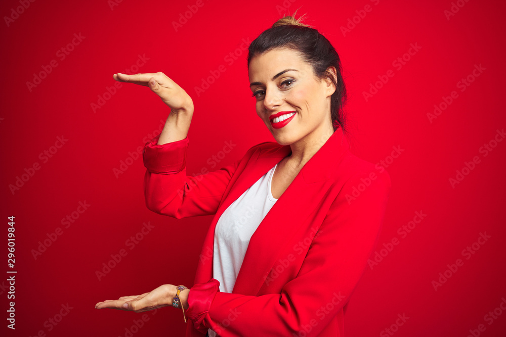 Young beautiful business woman standing over red isolated background gesturing with hands showing big and large size sign, measure symbol. Smiling looking at the camera. Measuring concept. - obrazy, fototapety, plakaty 