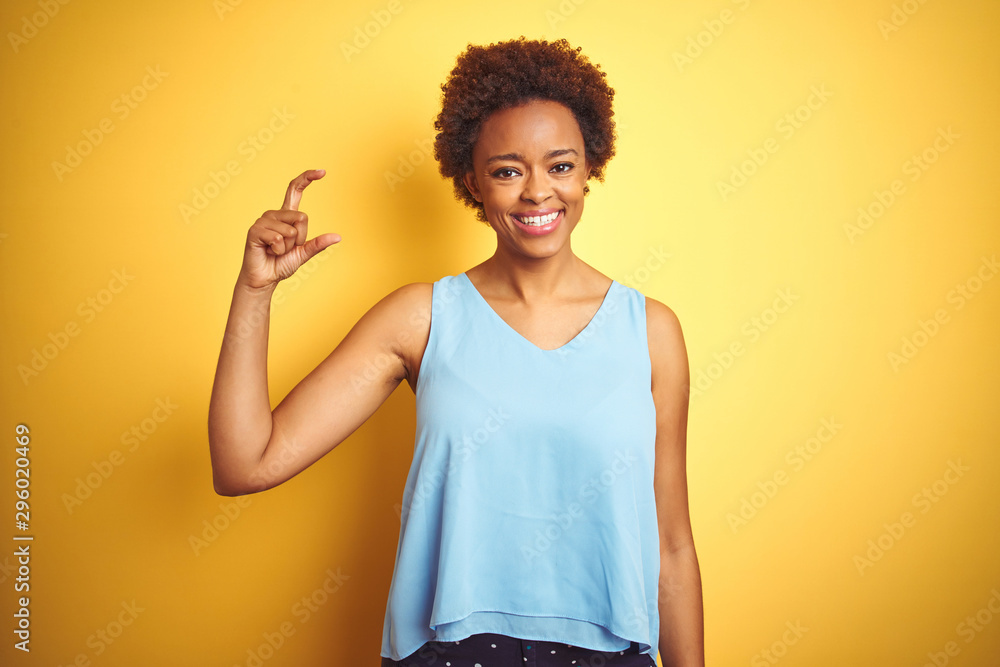 Beautiful african american woman wearing elegant shirt over isolated yellow background smiling and confident gesturing with hand doing small size sign with fingers looking and the camera Beautiful african american woman wearing elegant shirt over isolated yellow background smiling and confident gesturing with hand doing small size sign with fingers looking and the camera