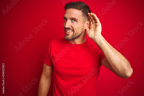 Young handsome man wearing casual t-shirt over red isolated background smiling with hand over ear listening an hearing to rumor or gossip. Deafness concept. © Krakenimages.com