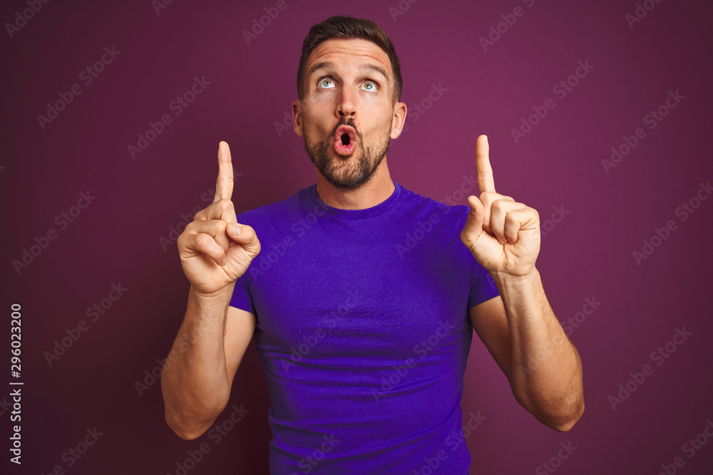 Young man wearing casual purple t-shirt over lilac isolated background amazed and surprised looking up and pointing with fingers and raised arms. Young man wearing casual purple t-shirt over lilac isolated background amazed and surprised looking up and pointing with fingers and raised arms.