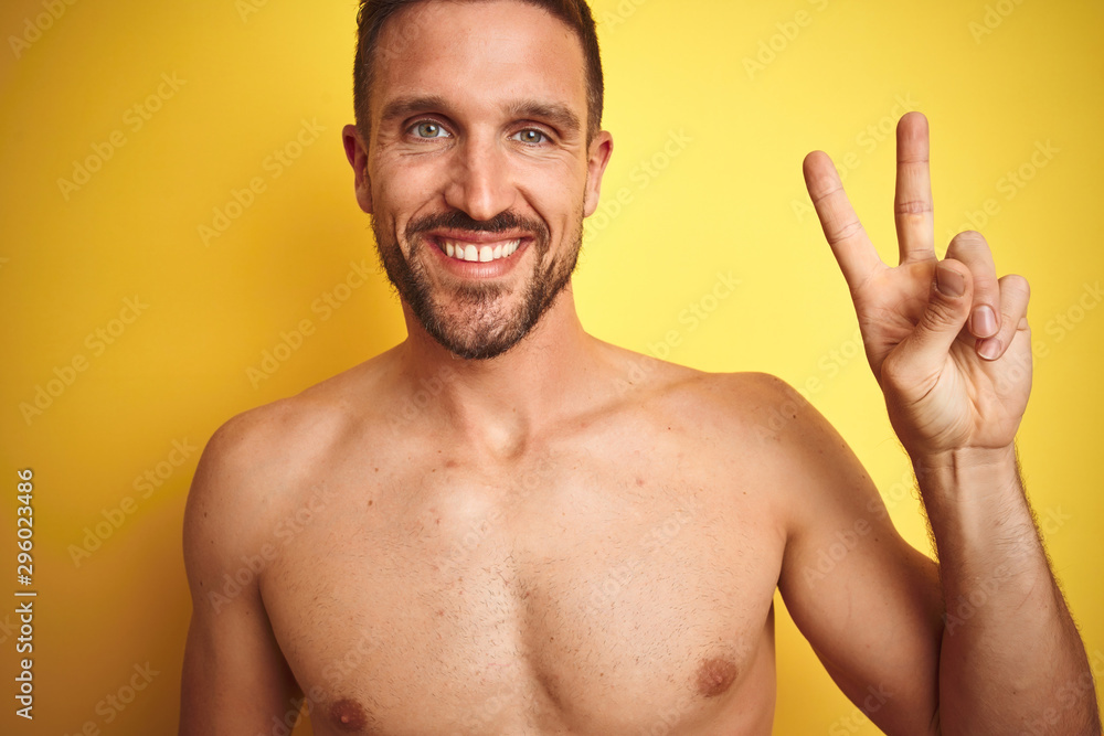Sexy young shirtless man showing nude fitness chest over yellow isolated background smiling with happy face winking at the camera doing victory sign. Number two. Sexy young shirtless man showing nude fitness chest over yellow isolated background smiling with happy face winking at the camera doing victory sign. Number two.