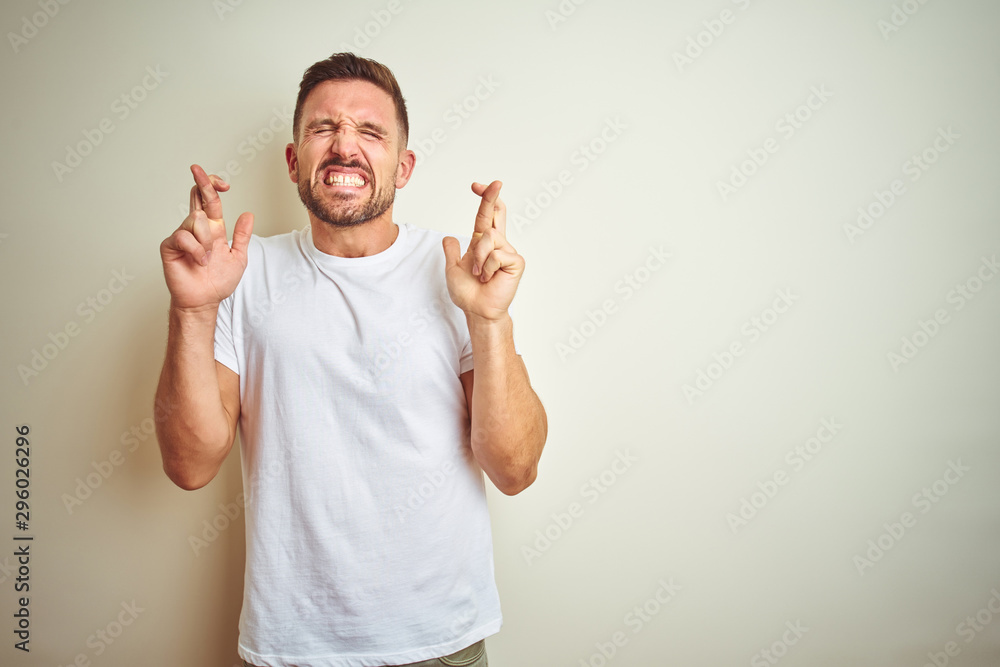 Young handsome man wearing casual white t-shirt over isolated background gesturing finger crossed smiling with hope and eyes closed. Luck and superstitious concept. Young handsome man wearing casual white t-shirt over isolated background gesturing finger crossed smiling with hope and eyes closed. Luck and superstitious concept.