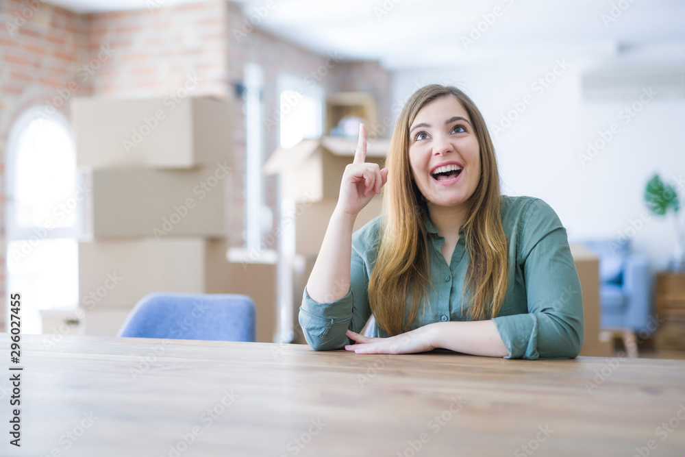 Young woman sitting on the table with cardboard boxes behind her moving to new home pointing finger up with successful idea. Exited and happy. Number one. Young woman sitting on the table with cardboard boxes behind her moving to new home pointing finger up with successful idea. Exited and happy. Number one.