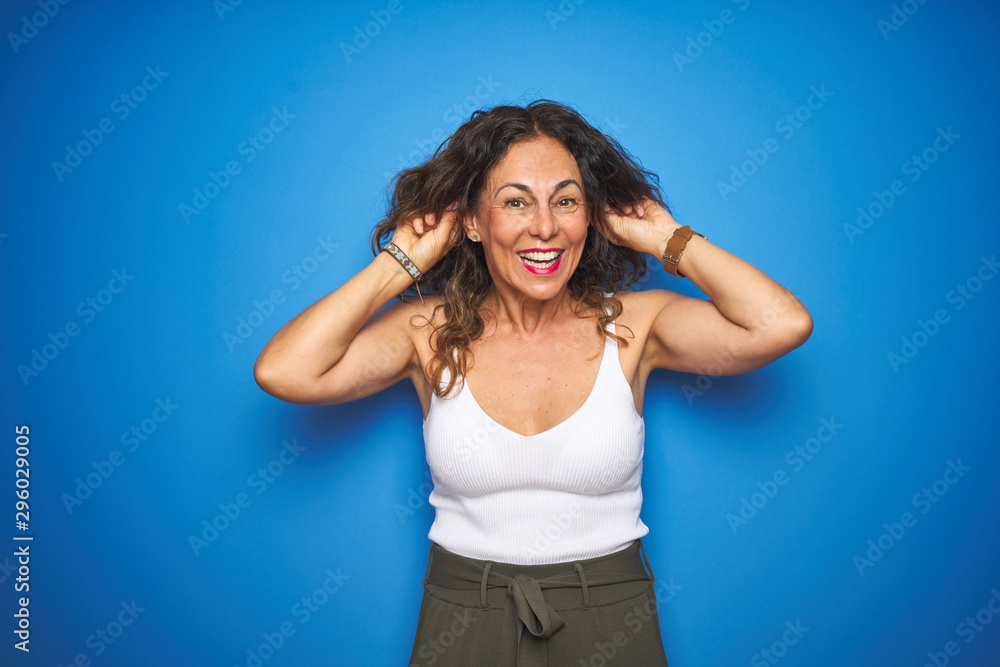 Middle age senior woman with curly hair standing over blue isolated background Smiling pulling ears with fingers, funny gesture. Audition problem Middle age senior woman with curly hair standing over blue isolated background Smiling pulling ears with fingers, funny gesture. Audition problem