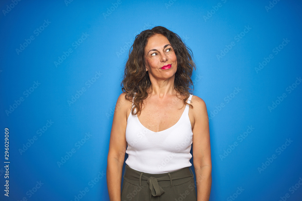 Middle age senior woman with curly hair standing over blue isolated background smiling looking to the side and staring away thinking.