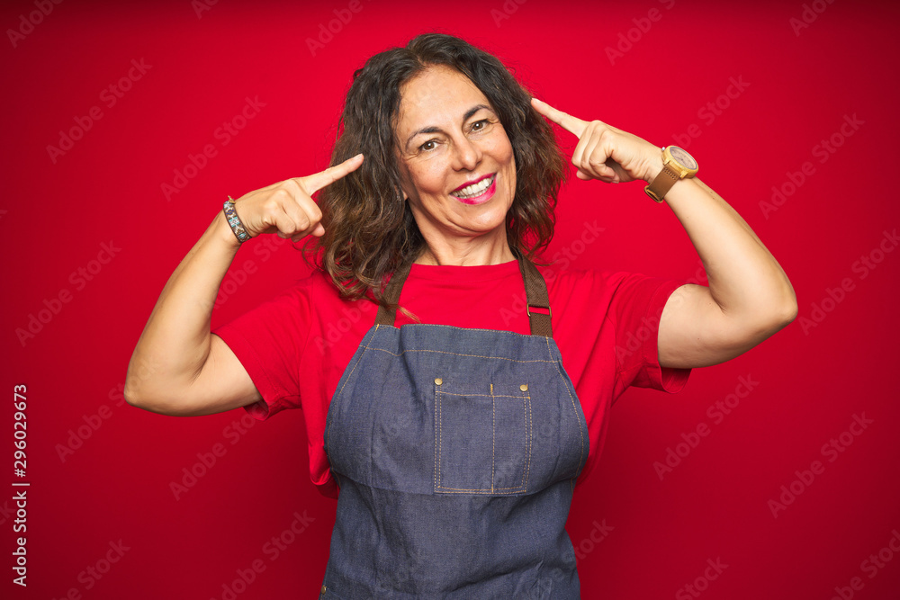 Middle age senior woman wearing apron uniform over red isolated background smiling pointing to head with both hands finger, great idea or thought, good memory Middle age senior woman wearing apron uniform over red isolated background smiling pointing to head with both hands finger, great idea or thought, good memory