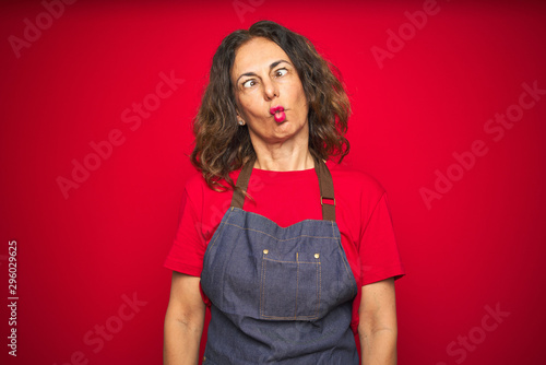 Vászonkép Middle age senior woman wearing apron uniform over red isolated background making fish face with lips, crazy and comical gesture