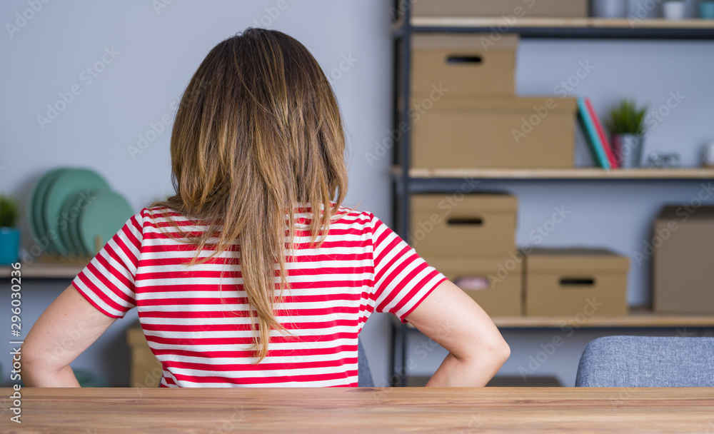 Middle age senior woman sitting at the table at home standing backwards looking away with arms on body Middle age senior woman sitting at the table at home standing backwards looking away with arms on body