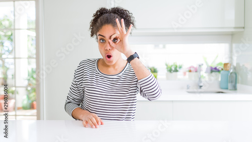 Beautiful african american woman with afro hair wearing casual striped sweater doing ok gesture shocked with surprised face  eye looking through fingers. Unbelieving expression.
