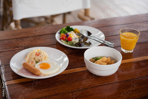 Breakfast includes bread  fried egg  various vegetable salads  sausages  fried rice  and orange juice on a brown wooden table