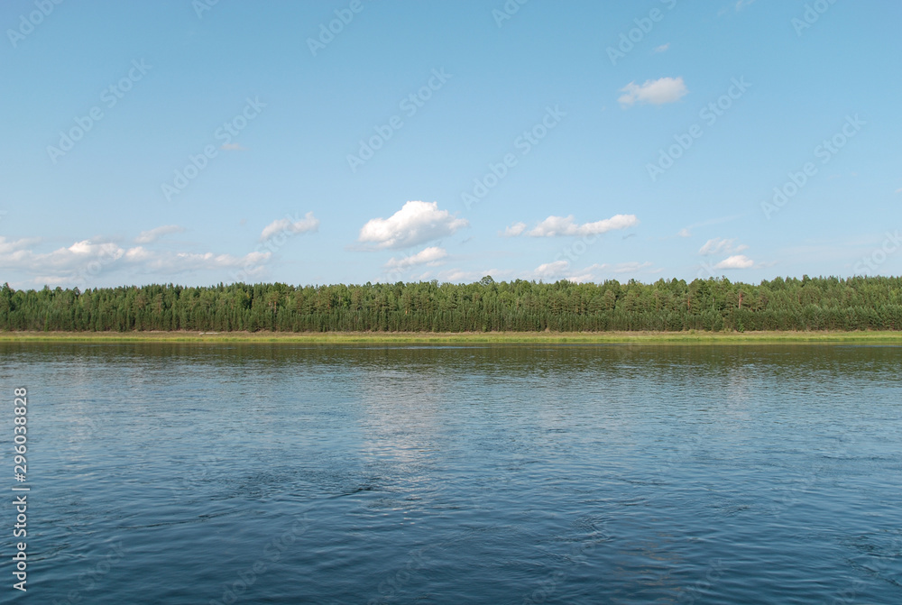A wide river with a green shore, on which there are numerous trees and fields. A clear sunny day in summer.