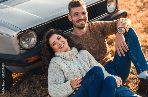 Beautiful young couple enjoying time together outdoor sitting on meadow leaning on old fashioned car embracing each other.