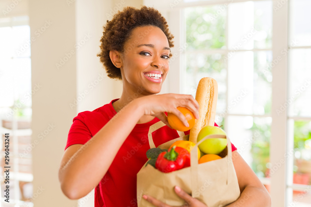 Young beautiful african american woman holding paper bag full of fresh healthy groceries and picking vegetables Young beautiful african american woman holding paper bag full of fresh healthy groceries and picking vegetables