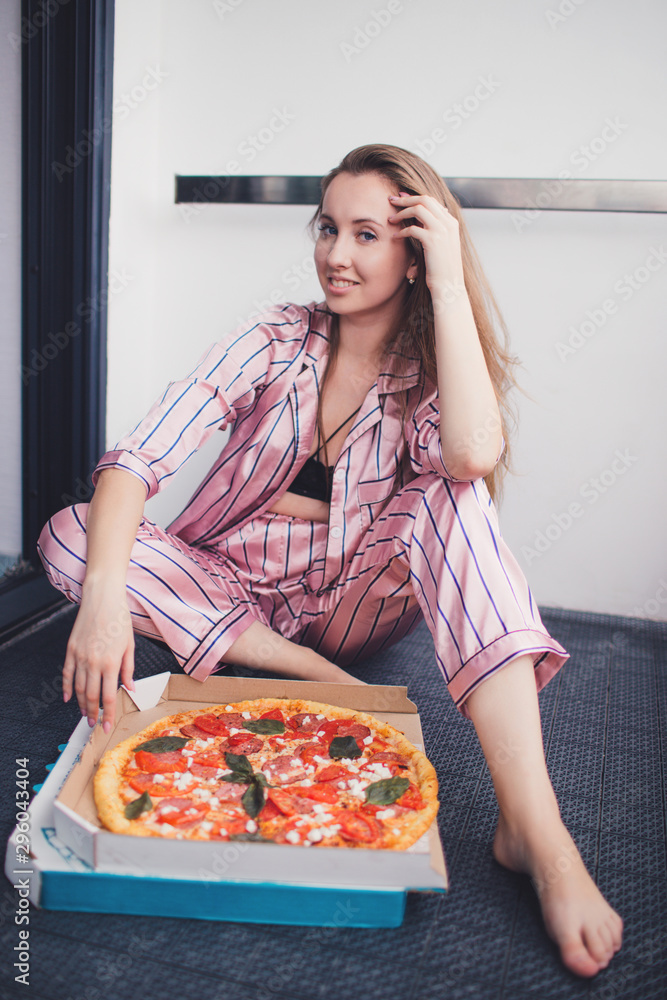 Young woman eating a piece of pizza sitting on floor wearing pajamas. Birthday party. Bachelorette party. Christmas party.