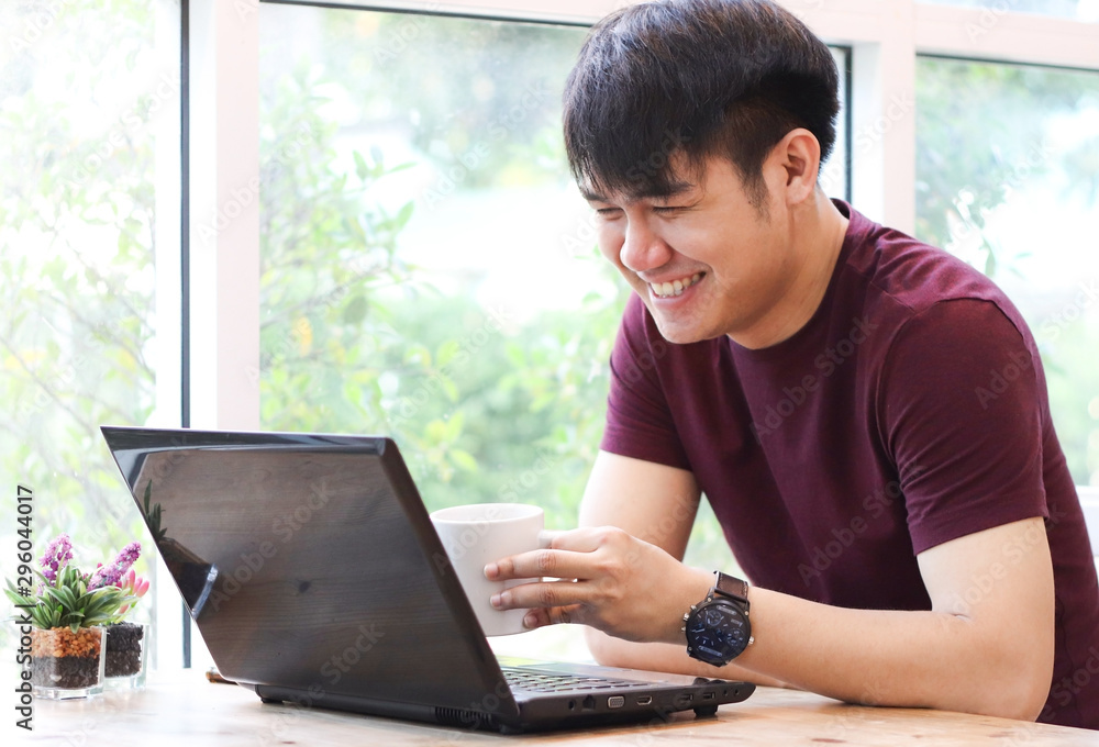 Asian young man sitting in coffee shop with computer laptop on the table , looking at computer screen and laughing. Asian young man sitting in coffee shop with computer laptop on the table , looking at computer screen and laughing.
