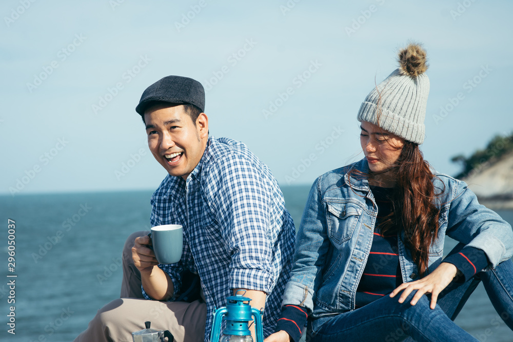 Happy Asian couple holding coffee cups on mountain peak together.