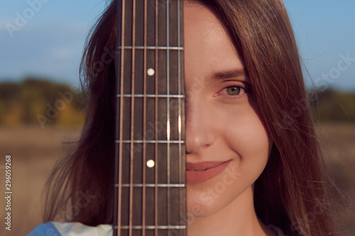 Closeup of a beautiful smiling girls face with guitar neck. Young musician with musical instrument. Caucasian girl with gorgeous eyes. Sunny day.