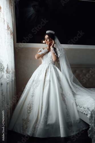Fashion photo of beautiful bride with dark hair in elegant wedding dress and crown posing in room in the wedding morning. Beautiful bride with professional make up and hairstyle.
