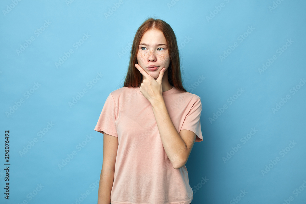 Pensive ginger girl holding her chin and looking aside over blue background, let me think, close up portrait, girl making decision Pensive ginger girl holding her chin and looking aside over blue background, let me think, close up portrait, girl making decision